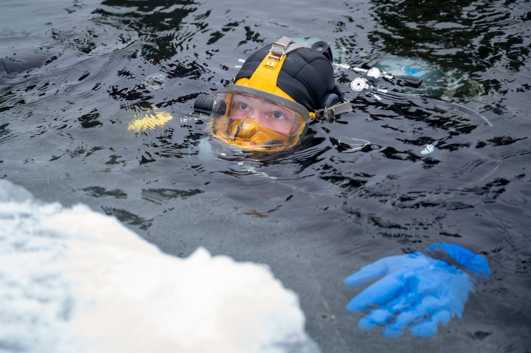 A sailor wearing a wet suit and diving gear holds their head above Artic water with snow in the left foreground.