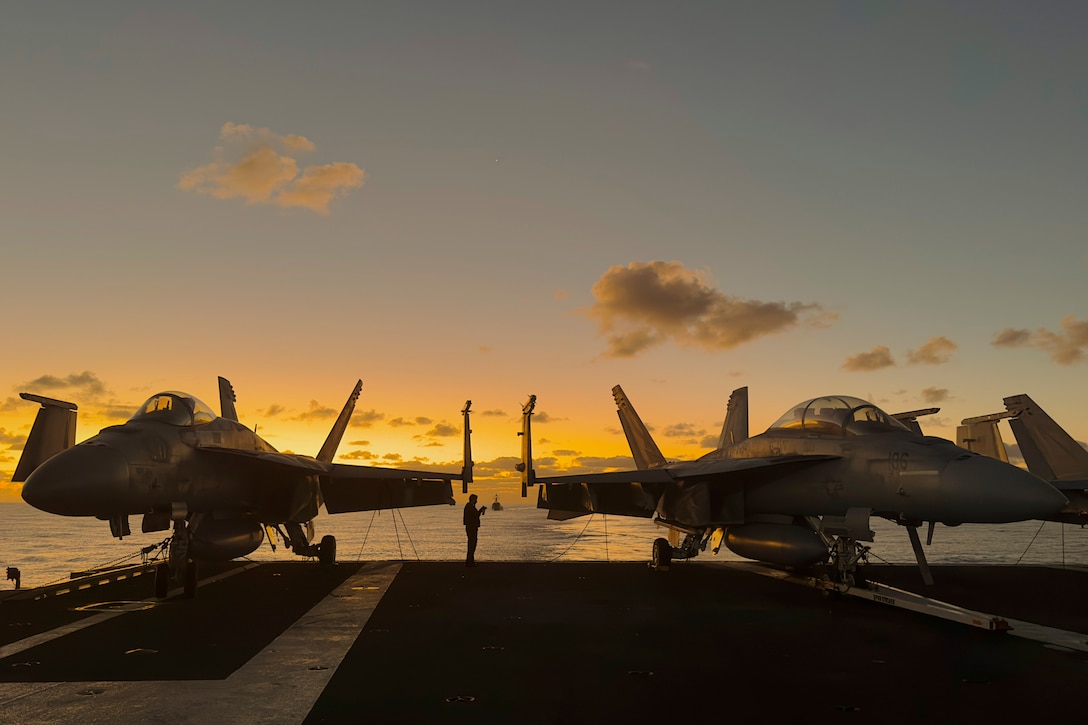 A sailor stands in between two aircraft parked on the edge of a ship as another ship sails in the background under a sunlit sky.