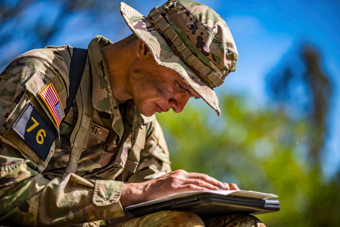 A soldier sits while plotting points on a paper with blurred trees in the background.