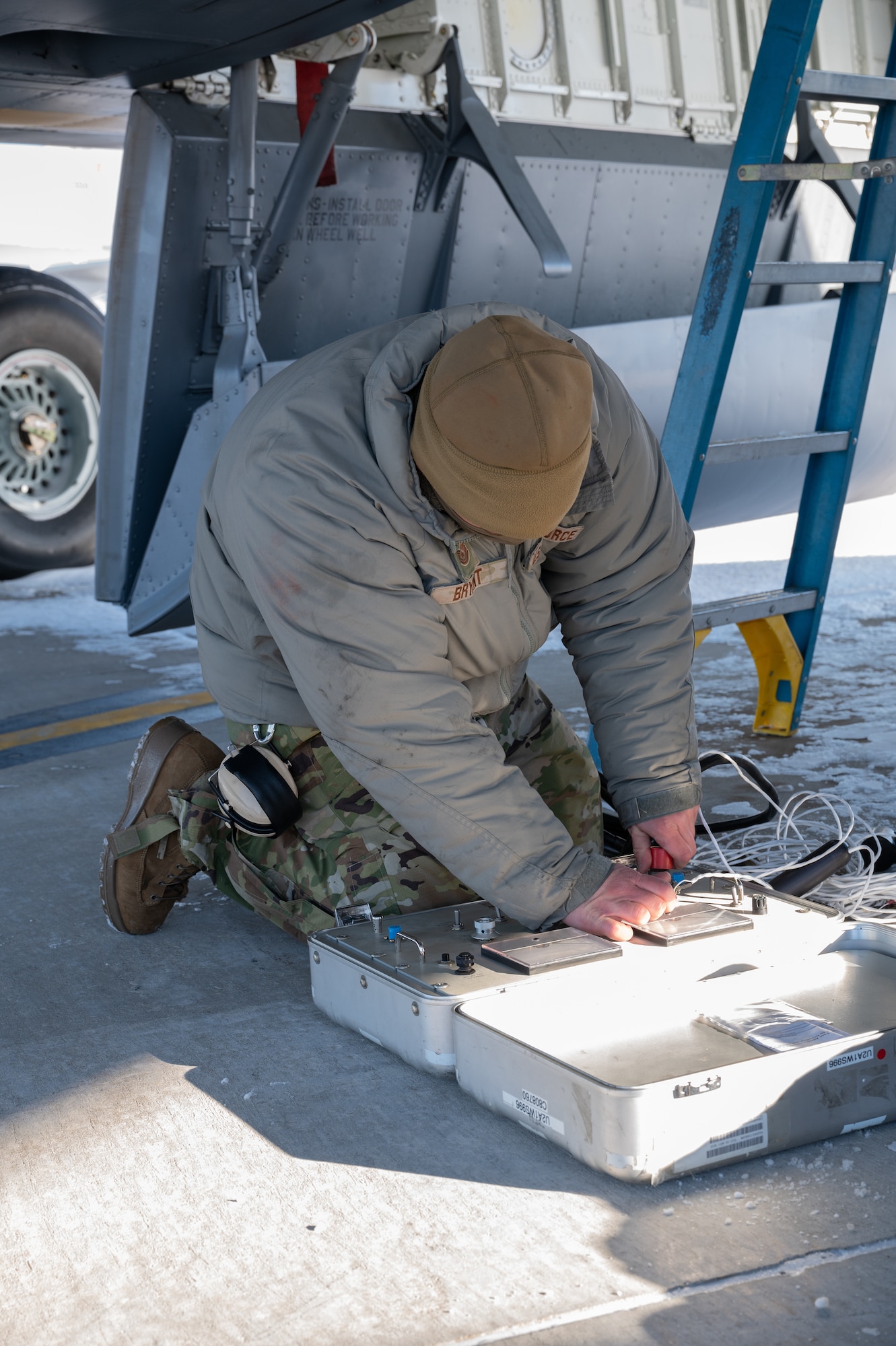 Adrian Bryant, an Air Force electrical technician, is kneeling on the ground and working within a white, brief-case style box. He is positioned to the side of a blue ladder and is wearing cold weather Air Force-issued gear.