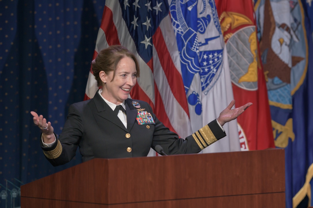 A Navy Vice Admiral gives a speech at a podium with military flags in the background.