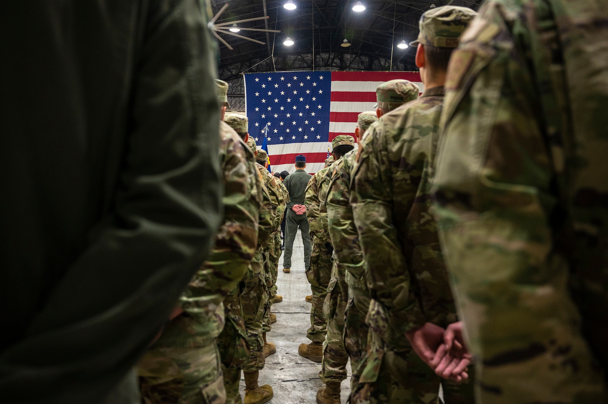 U.S. service members stand in formation, appearing from behind at below shoulder height, with a large U.S. flag in the backdrop.