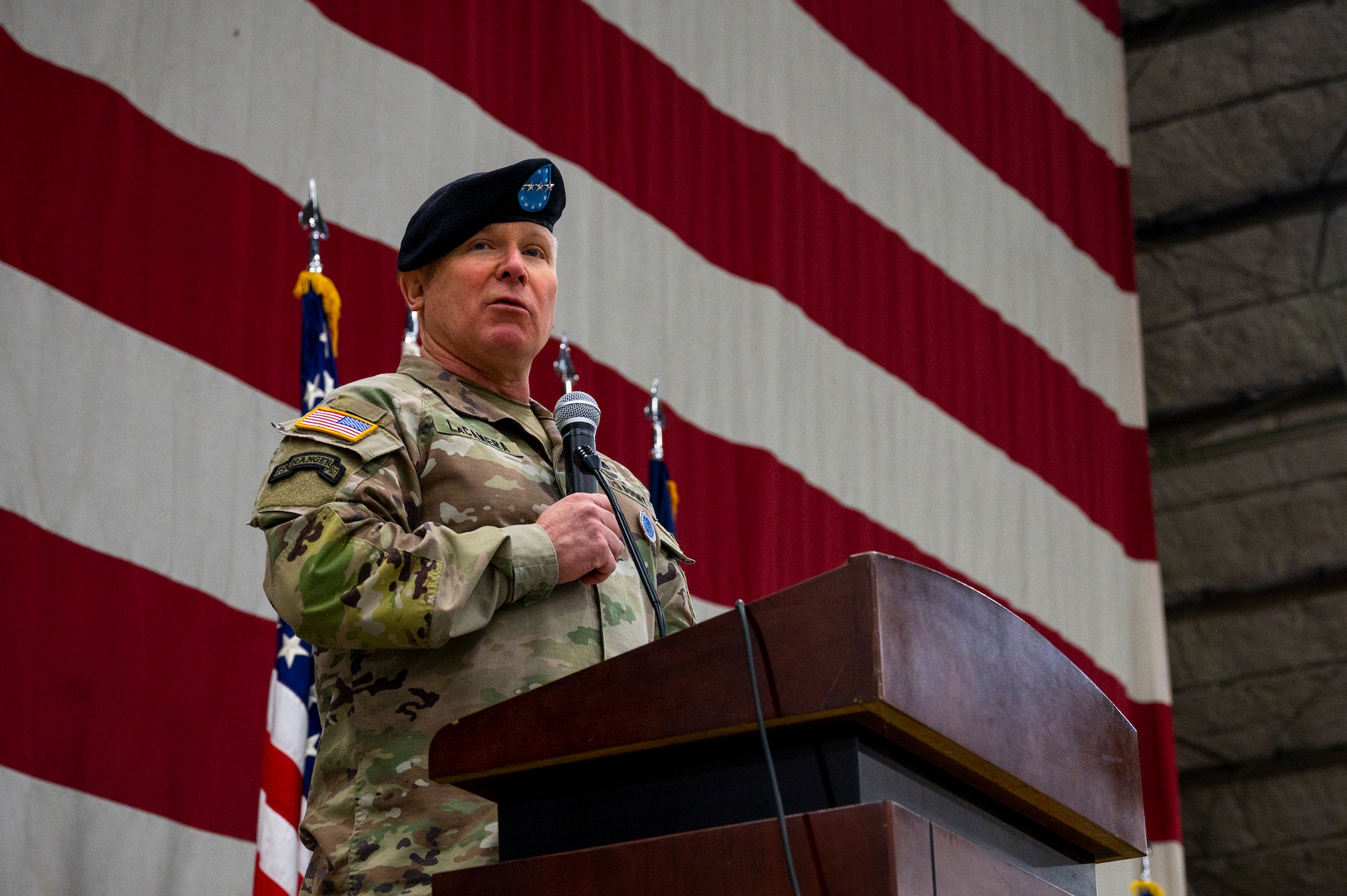 U.S. Army Gen. Paul LaCamera holds a microphone while speaking behind a podium in front of a large U.S. flag backdrop.