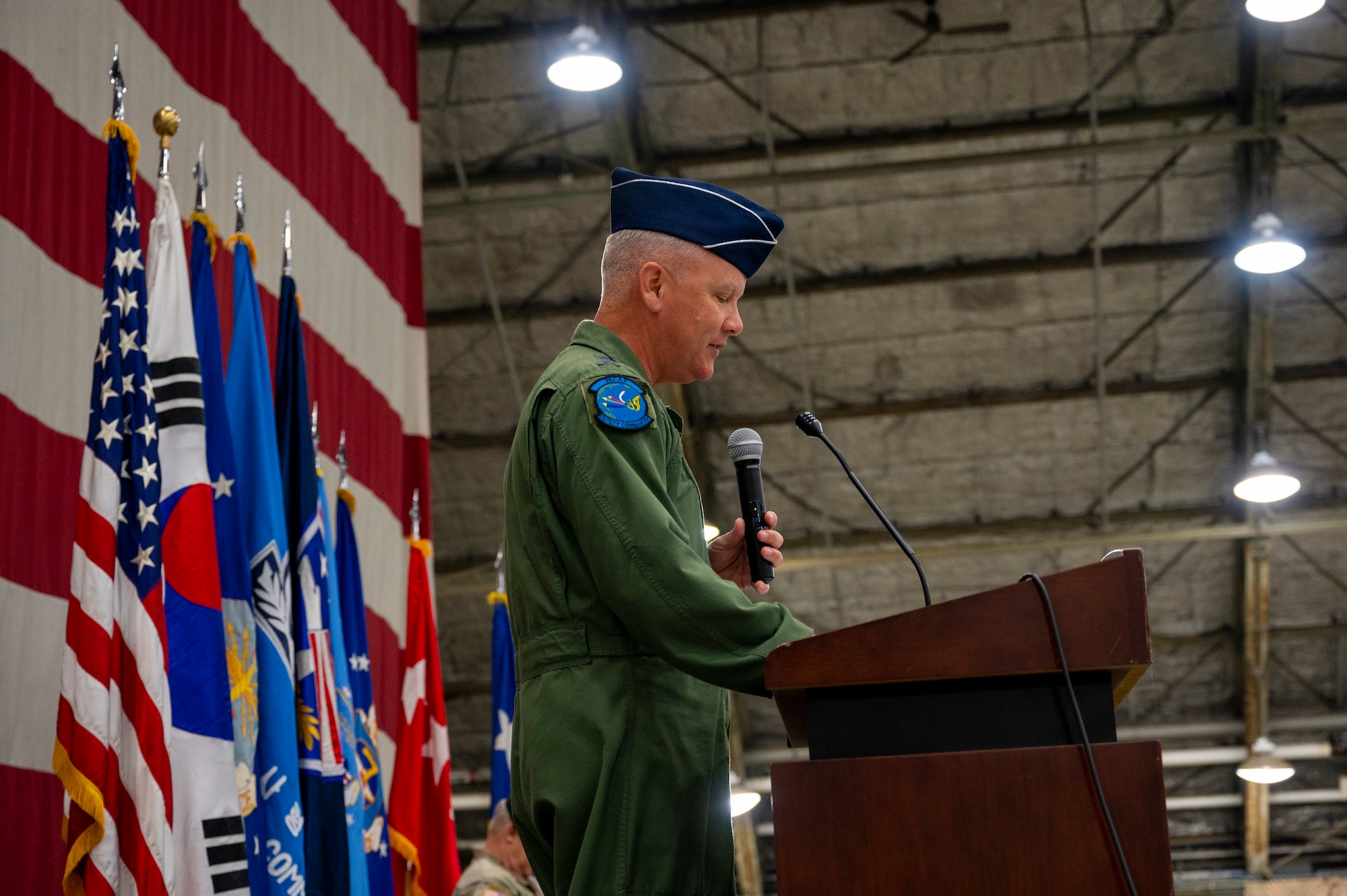 Lt. Gen. James Jacobson, Headquarters Pacific Air Forces deputy commander, holds a microphone from behind a podium while on a stage with a U.S. flag backdrop.