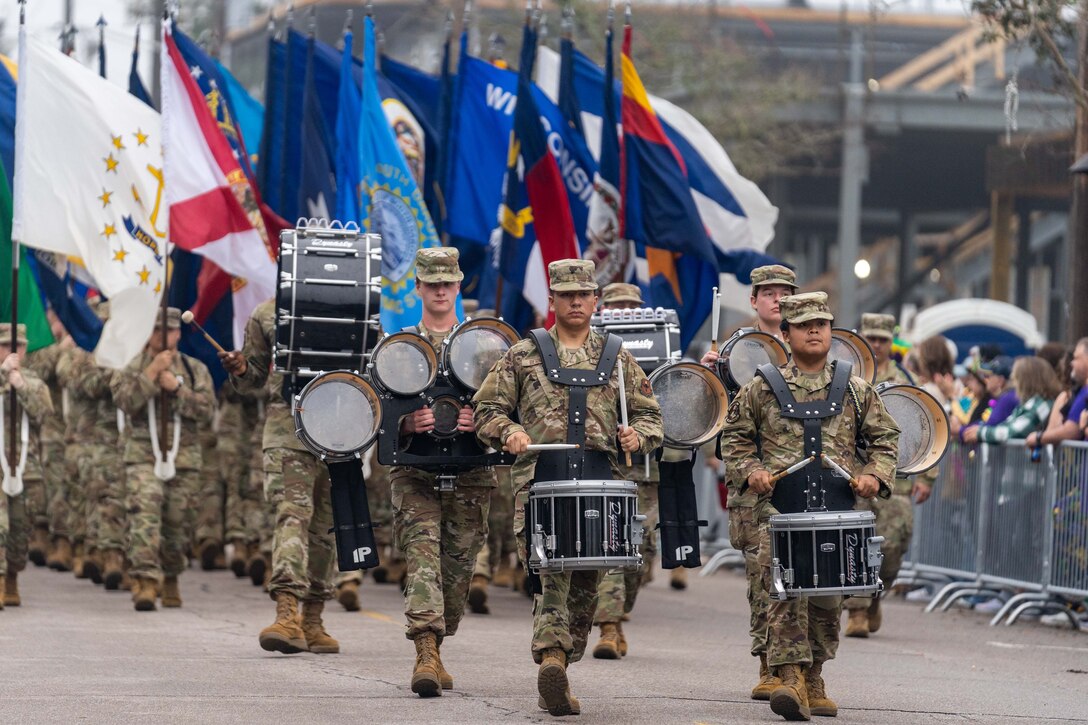 Airmen playing different types of drums lead a parade with many flags as spectators watch from behind barriers.