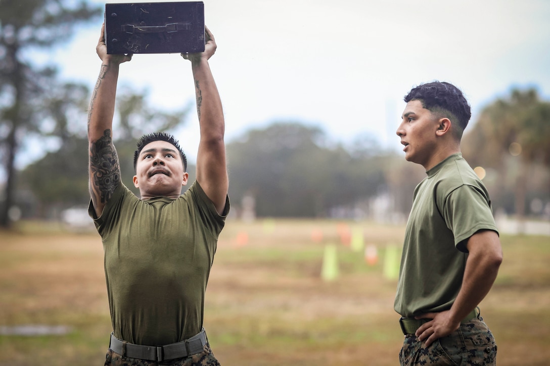 A Marine holds an object over their head as a fellow Marine watches as they stand in a field.