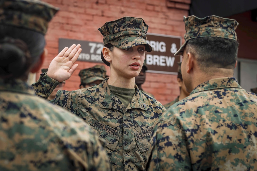A Marine raises their right hand while facing two Marines as others stand in the background in formation.