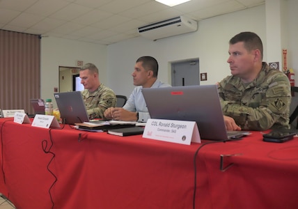 2 Army officers and a civilian seated at a table with red tablecloth.