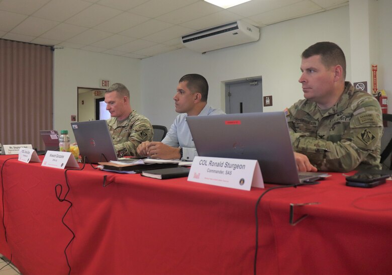 2 Army officers and a civilian seated at a table with red tablecloth.