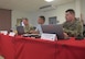 2 Army officers and a civilian seated at a table with red tablecloth.