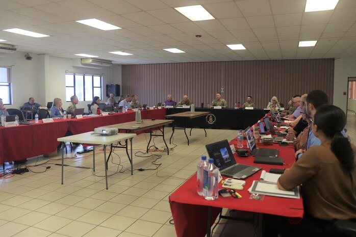 Conference room with US Army officers and civilians seated at table covered in red table cloth.