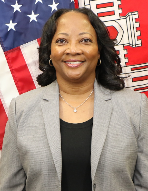 An smiling African American woman, Yvonne Prettyman-Beck, stands up wearing a grey jacket and a black shirt, in front of a red, white and blue, U.S. Flag and the red flag with a white castle representing the United Stated Army Corps of Engineers.