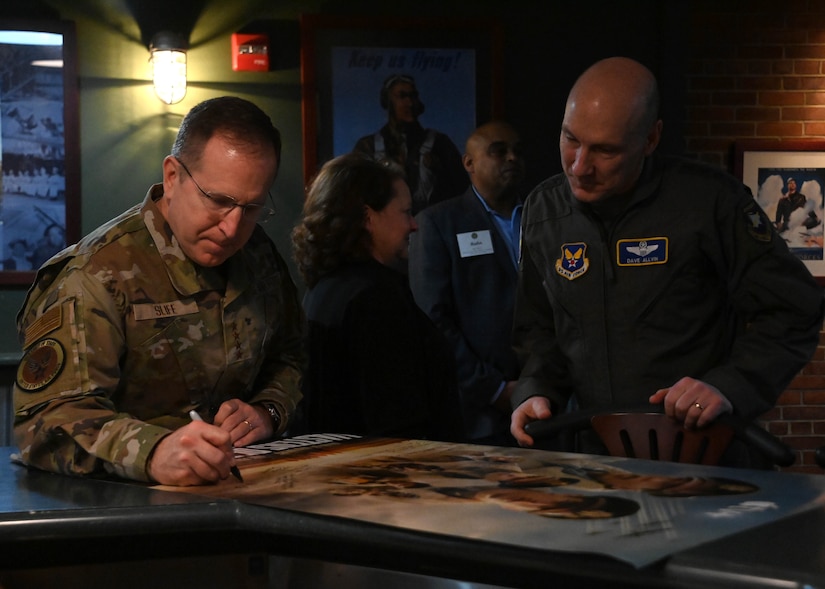 Two men stand by one another taking turns signing a poster.