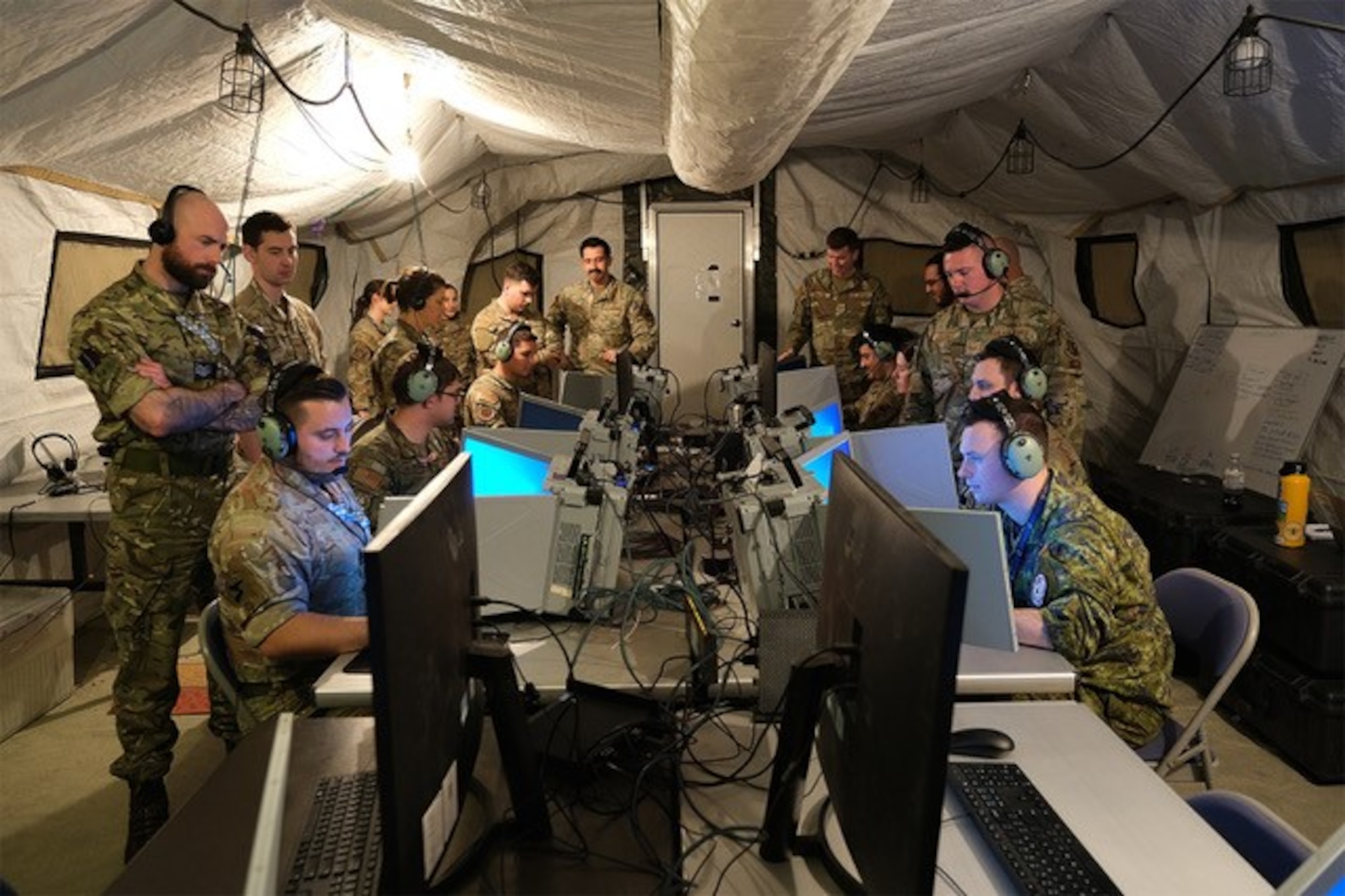 photo of uniformed military members working in a tent, some sitting at mobile equipment with headphones while others stand behind them watching