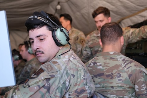 photo of uniformed military members working in a tent, some sitting at mobile equipment with headphones while others stand behind them watching