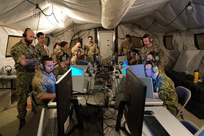 photo of uniformed military members working in a tent, some sitting at mobile equipment with headphones while others stand behind them watching