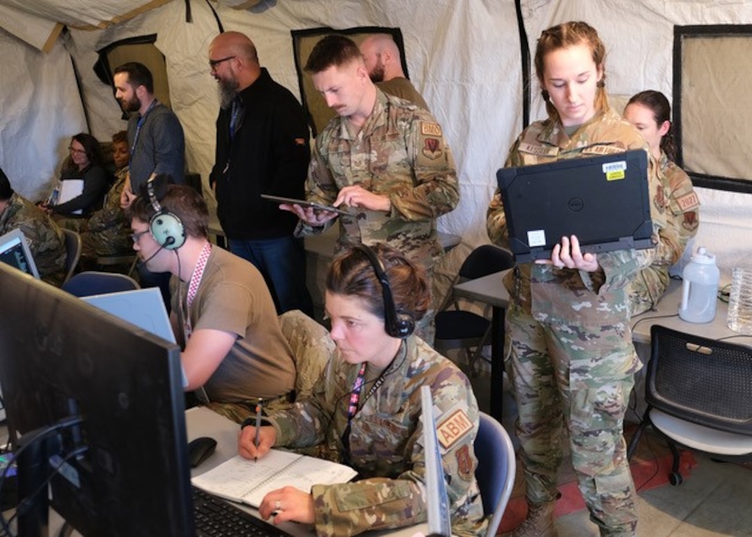 photo of uniformed military members working in a tent, some sitting at mobile equipment with headphones while others stand behind them watching