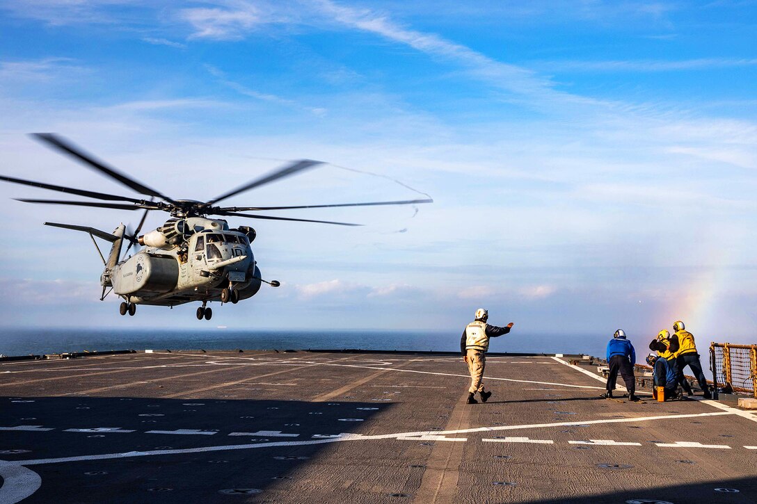 A partial rainbow appears behind sailors on the  far edge of the deck of a ship observing a helicopter about to land.