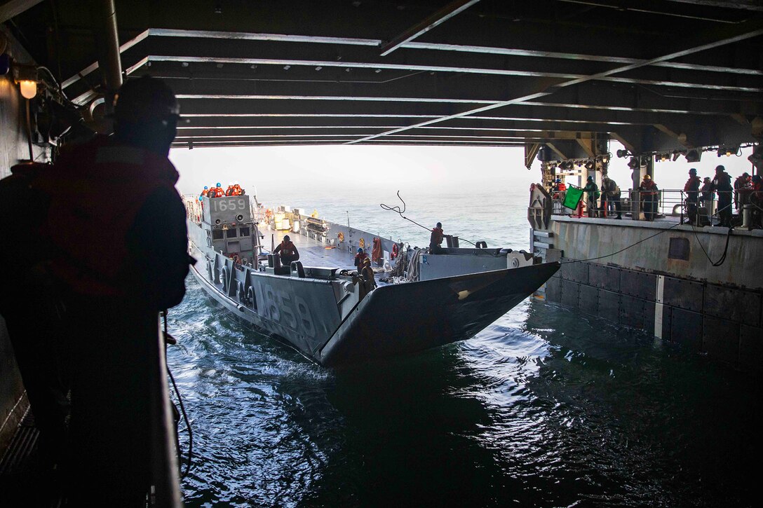 A landing craft utility vehicle with a few sailors on board, enters the well deck of a ship as other sailors look on from the sidelines.