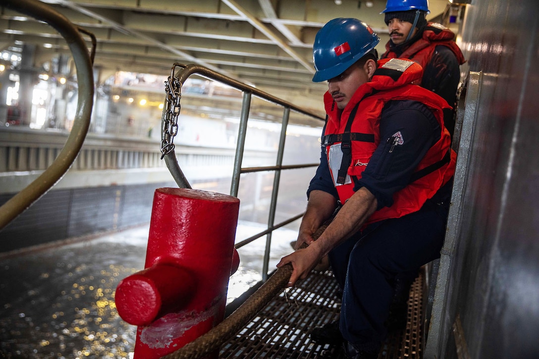 One of two sailors pulls on a rope; both wearing life vests and blue hard hats in the well deck of a ship.