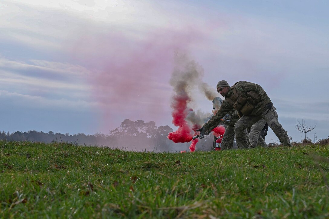 A group of airmen toss grenades emitting pink smoke while training in a large field.