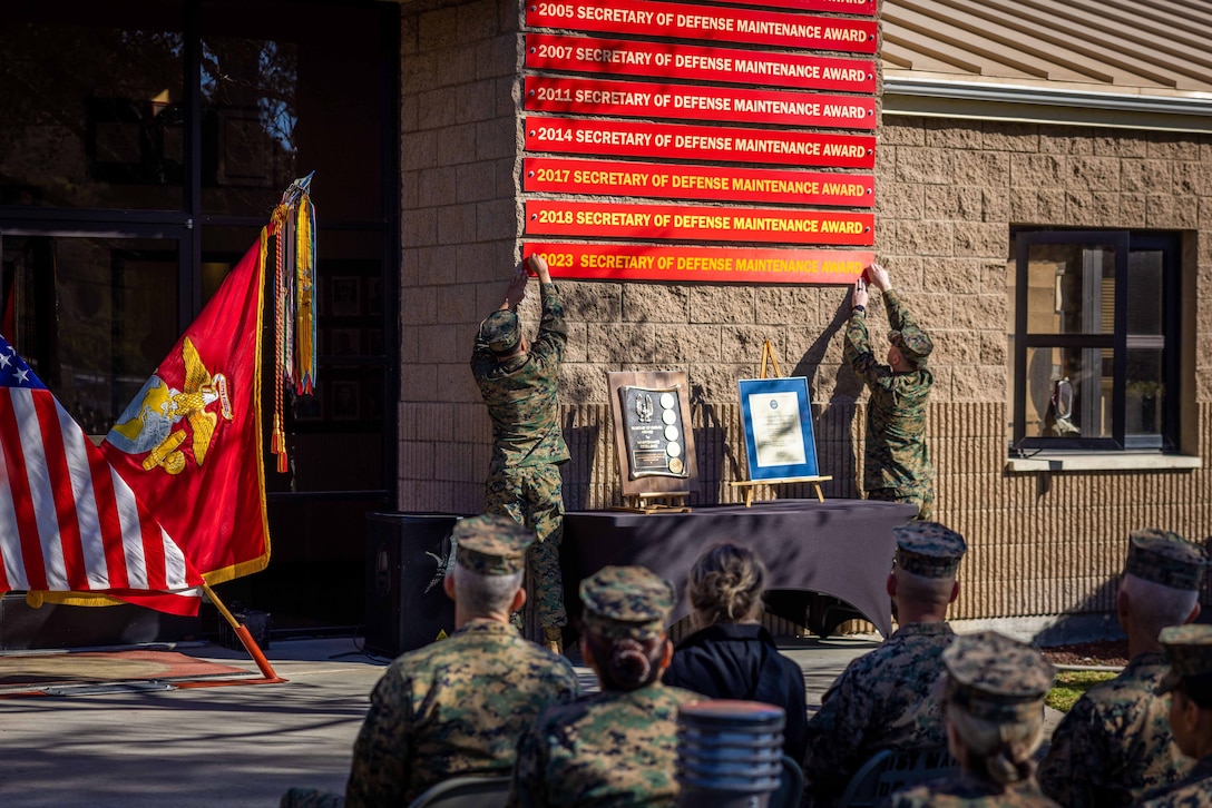 Two Marines attach a banner to a wall while other Marines sit in chairs and watch.