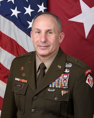 A man in an Army uniform poses in front of the American flag and the one-star general officer flag