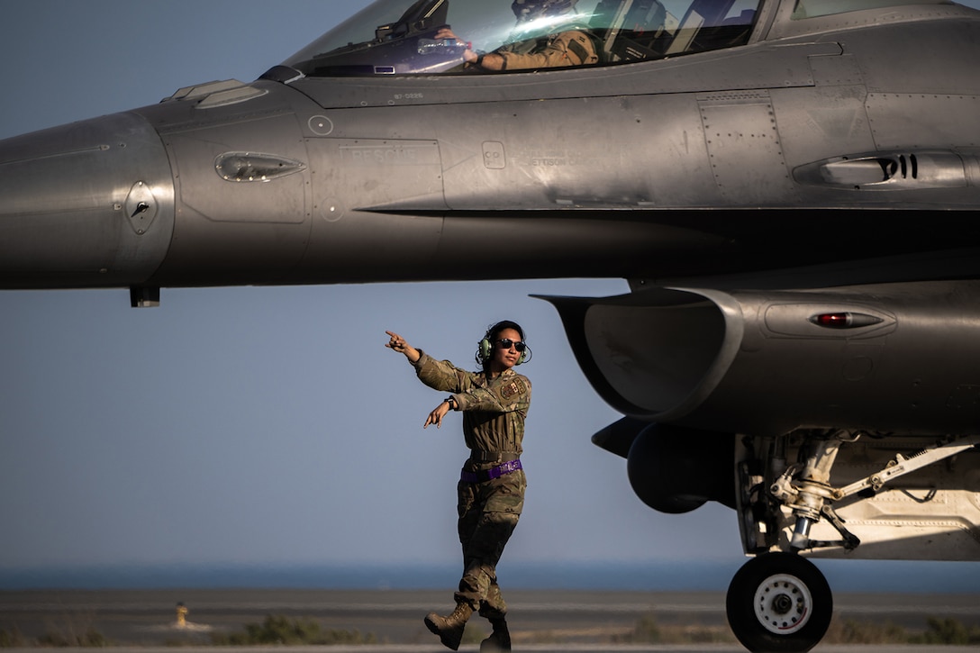 A uniformed airman, wearing a headset, points to direct a military aircraft before takeoff.