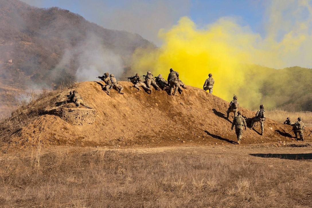 Soldiers fire weapons towards a mountain while laying on a dirt hill as clouds of yellow smoke fill the air.