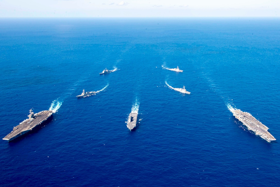 Seven ships sail in formation in a body of water as seen from above.