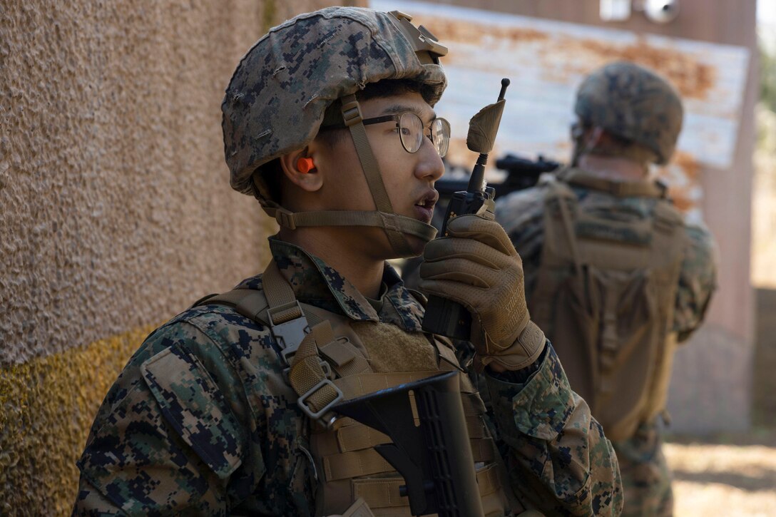 A Marine in tactical gear speaks on a handheld radio as a fellow Marine aims a weapon in the background.