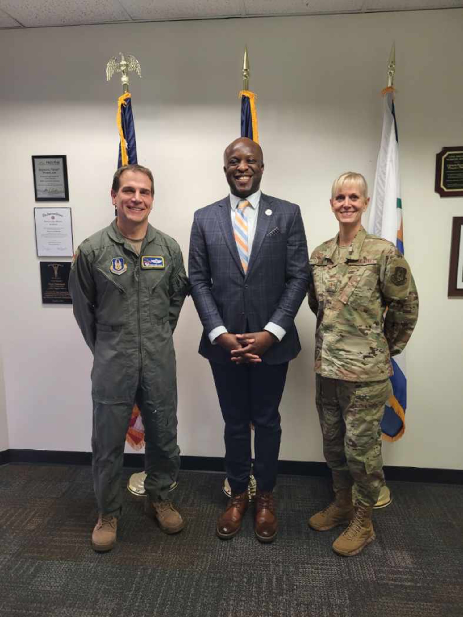 A man in a flight suit (left), a man in a business suit (middle), and a woman in operational Air Force duty uniform (right), stand next to each other with flags behind them.