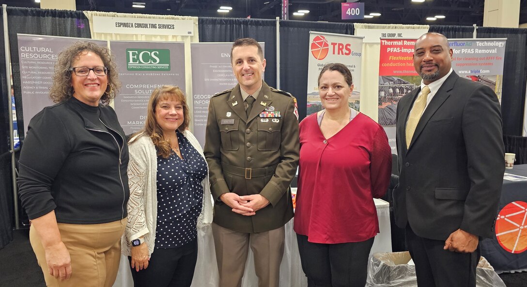 Groups of people from U.S. Army Corps of Engineers stand together to pose for a photo in business wear with booths surrounding them at the Small Business conference.