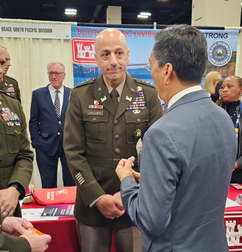 Groups of people from U.S. Army Corps of Engineers stand together to pose for a photo in business wear with booths surrounding them at the Small Business conference.