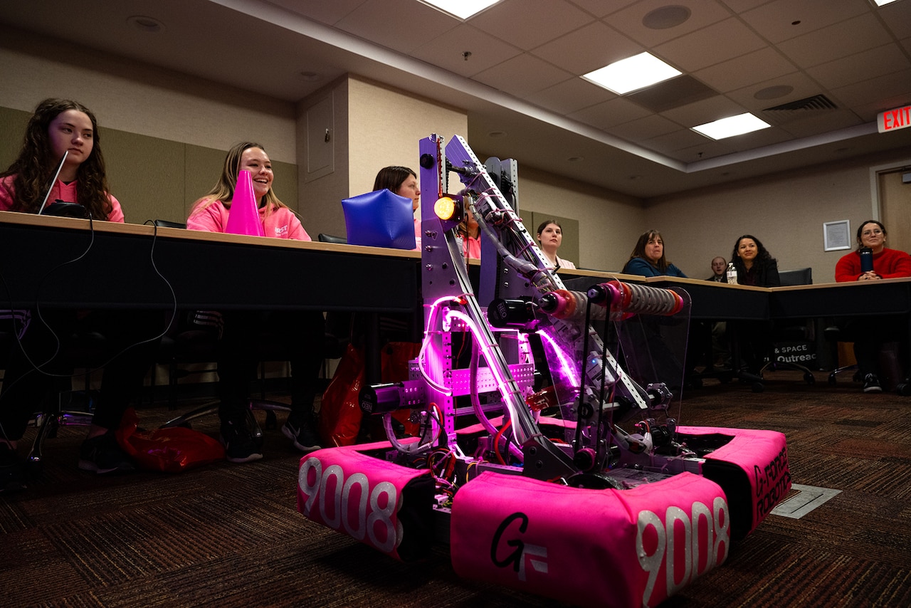 A robot sits on the floor in front of a table with several seated people.