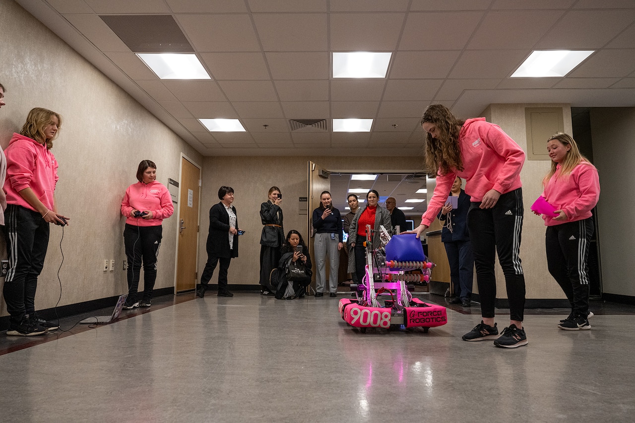 Two young people operate a robot while another places an object onto a robot.