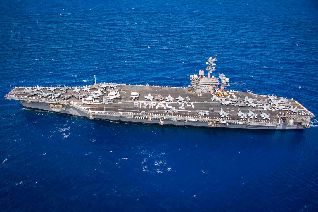 Oblique overhead view of a ship in blue waters, with sailors on the deck positioning themselves to spell "RIMPAC 24."