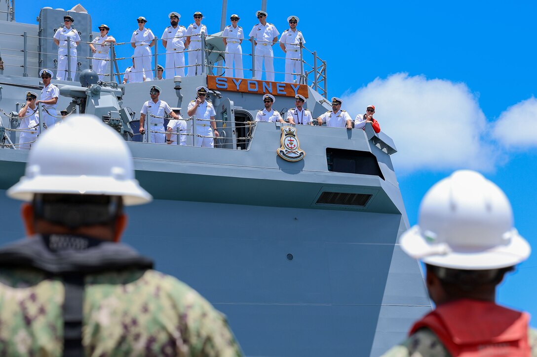 Two sailors in hard hats, shown from behind  at a dock, look up at a nearby ship with sailors manning the rails.