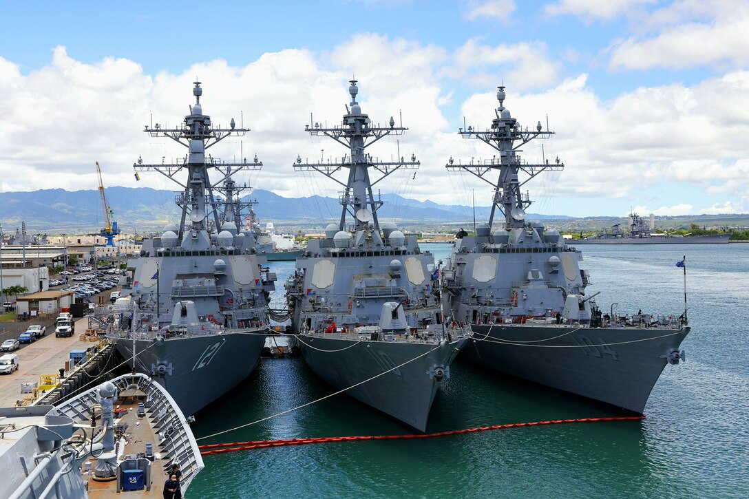 Three identical-looking ships are moored side-by-side at a dock.