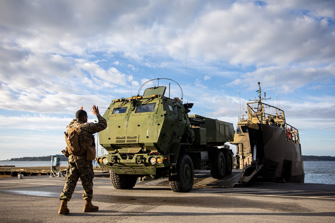 A Marine holds their arms up to direct an artillery vehicle onto the deck of a ship during daylight.