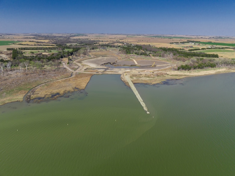 An aerial view of a lake. Green water in the foreground with brown and green land and blue sky in the background.