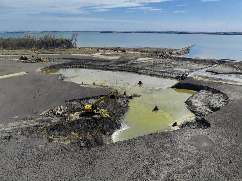 Two yellow pieces of heavy equipment sit on black dirt with a green pool of water to the right and blue water behind.