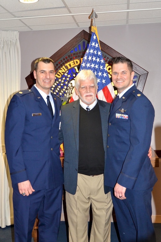 Three people stand in front of a flag and smile for the camera.