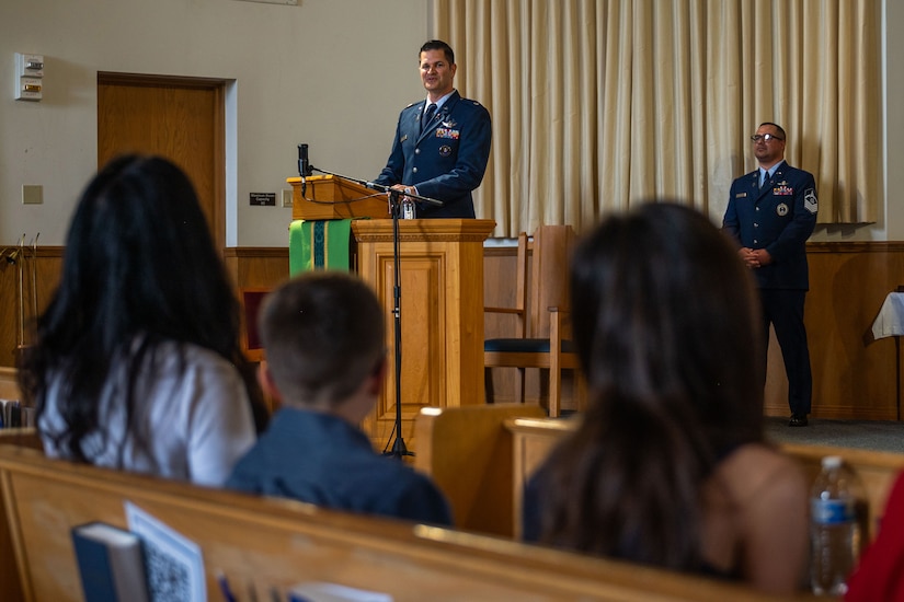 A military officer speaks to an audience in from a lectern in a chapel while another service members stands in the background.