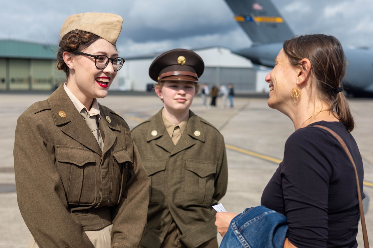 Two young people dressed in vintage military uniforms talk to a person whose back is toward the camera.