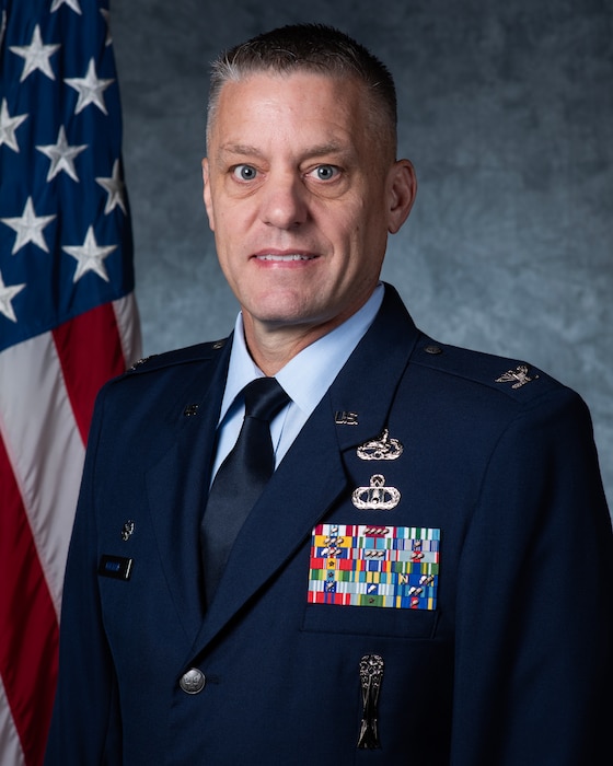 An Air Force Colonel in dress blues looks forward in front of an American flag and a grey background.