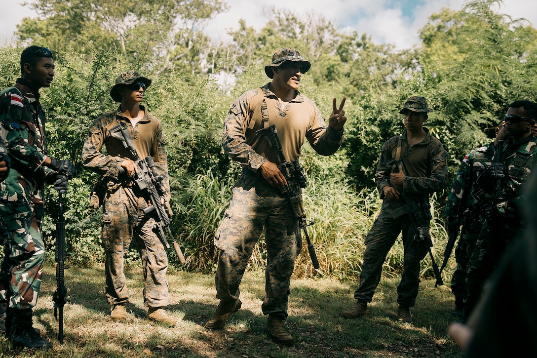 A U.S. Marine talks to several U.S. and foreign Marines standing in a woodsy area holding weapons.