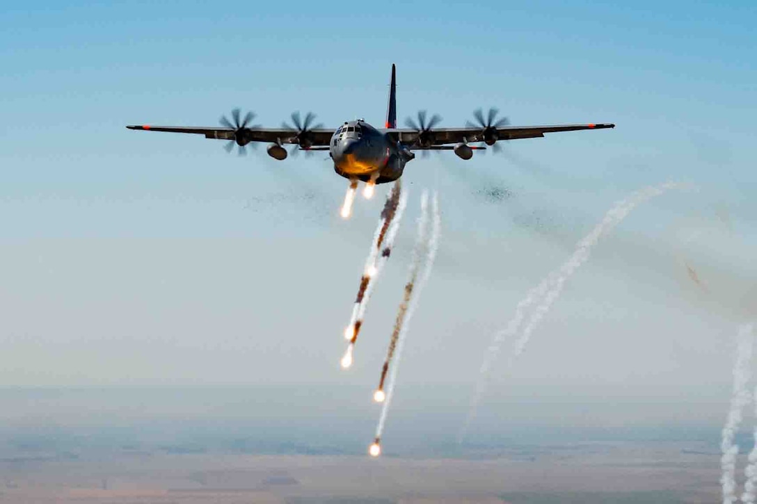 A large military aircraft releases chaff and flares as it flies across a light blue sky.