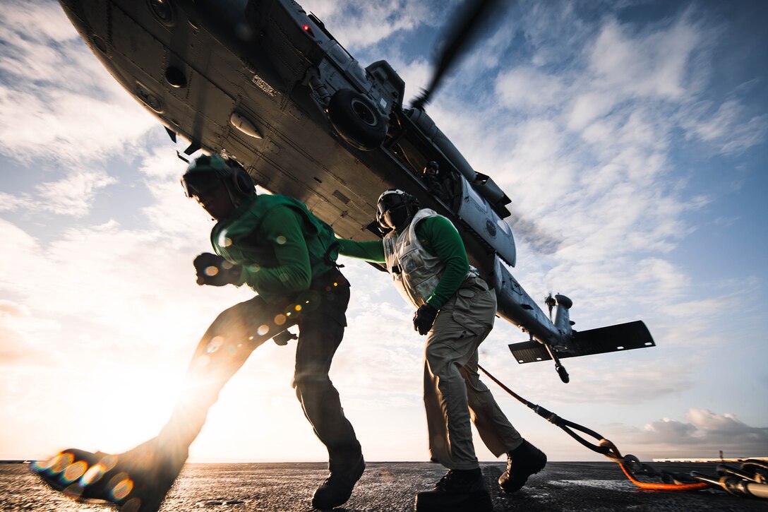 Service members work on the flight deck of a Navy ship as a helicopter drops nets from above.