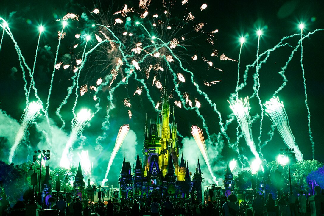 Green fireworks and vapor trails surround a castle structure at night as people watch.